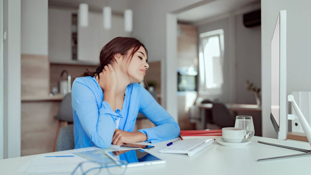 Person with long brown hair in a blue shirt rubs neck at desk, possibly needing a chiropractor in San Antonio.